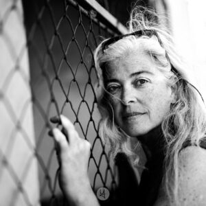 Black and white portrait of a woman (my wife) leaning on a wire-mesh fence. She grabbed with her hands the fence and is looking into the camera.