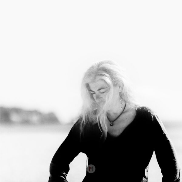 black and white high contrast portrait of my wife on a sunny day on the beach. The photo shows the upper part of her, she looks to the left corner of the picture, her hair is blowing gently in the wind. He background is blury, her shirt very dark so it gives a high contrast portrait.
