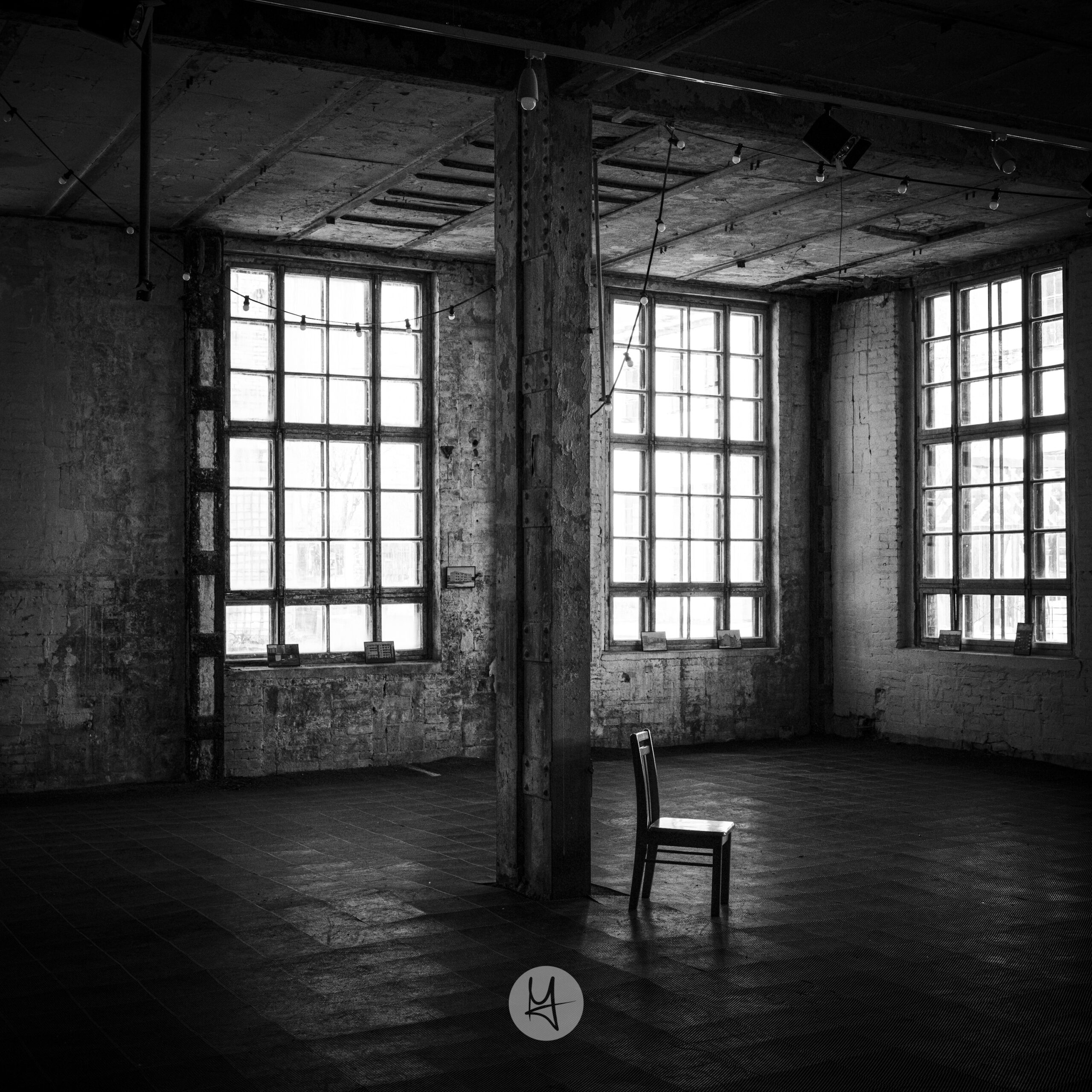 A high-contrast, black and white photograph of a large, empty, derelict room. A single wooden chair sits in the center foreground, its back facing a tall, weathered metal pillar. The room is brightly lit by natural light streaming through three large, multi-paned windows on the far wall. The floor is tiled, and the walls are made of exposed brick.
