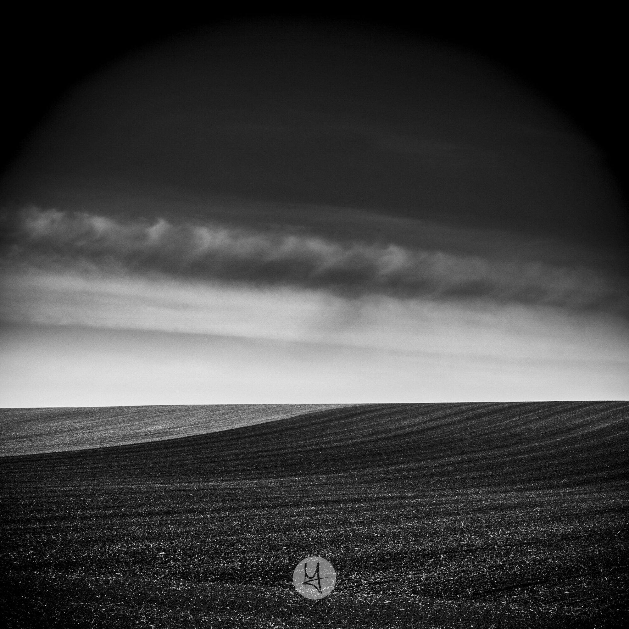 A black and white photo of a field. The foreground is a dark field with a faint pattern of furrows running from left to right, rising slightly to a curved horizon. A lighter band of sky sits above the horizon, with two distinct, dark cloud banks above it. The top of the frame is dark, with no detail visible.