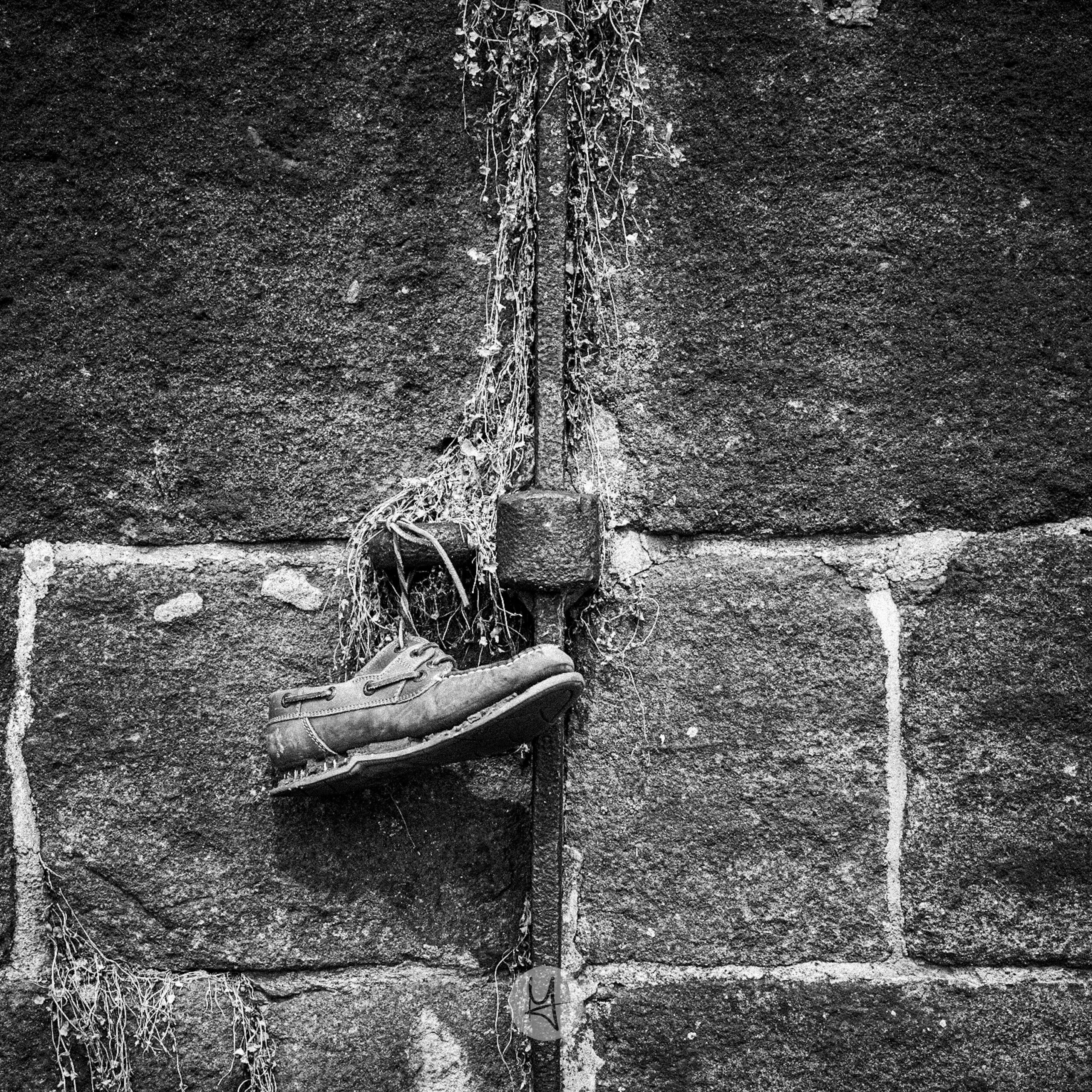 A black and white close-up photo of an old, worn boat shoe hanging from a metal rod on a stone wall. The shoe is suspended from the shoelaces and appears to be heavily weathered. The stone wall is made of rough-hewn blocks. A small vine with dead leaves hangs from the top of the frame, trailing down the metal rod and over the shoe.