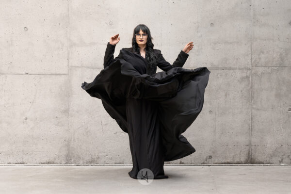A portrait of a person with long dark hair and glasses, wearing a black top and a long black skirt. The skirt is caught in motion, billowing outwards. The person is standing in front of a concrete wall.