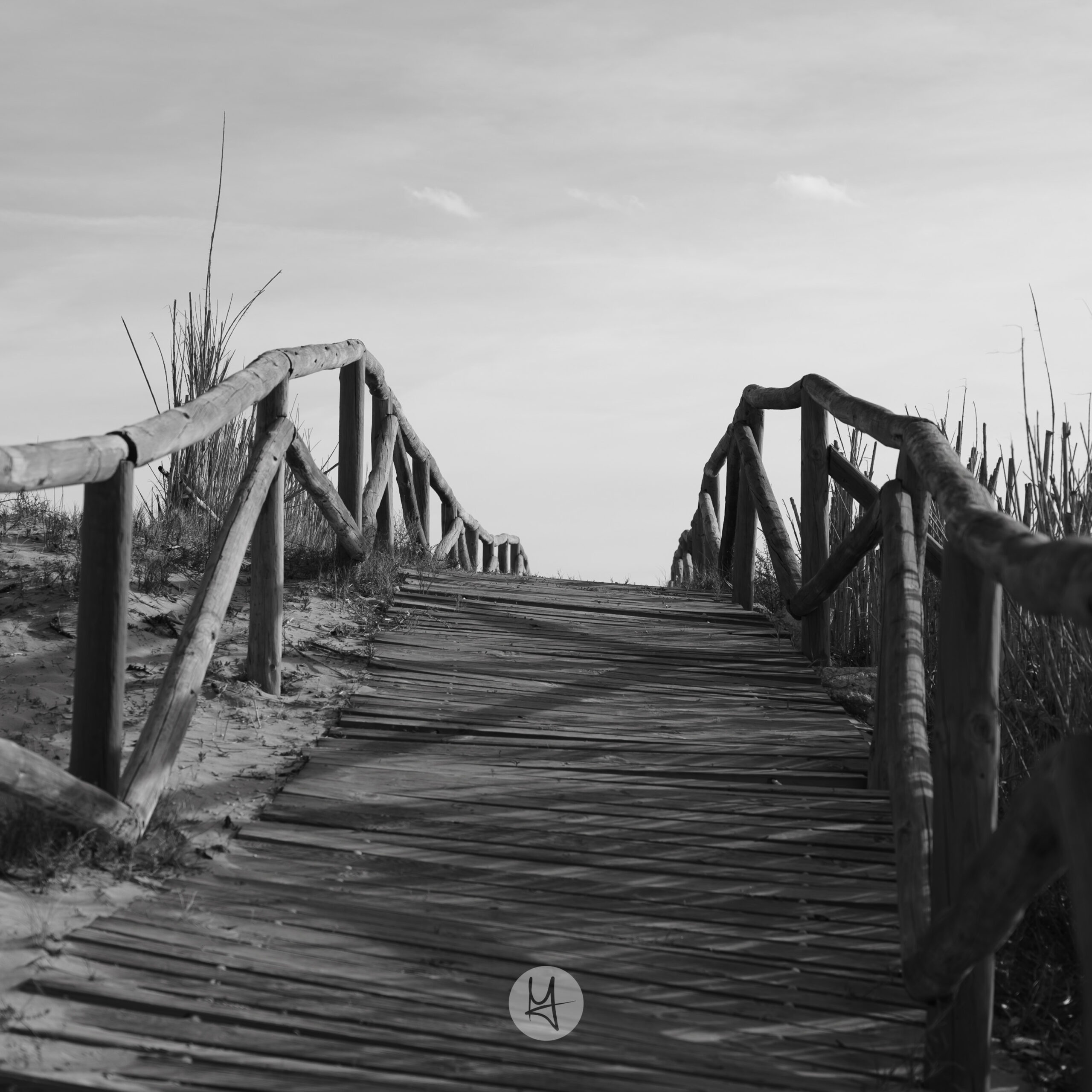 A black and white photo of a wooden path in the dunes at the coast side somewhere in Spain. The path is leading upwards so that you can’t see where it’s leading to. The sky is filled with a few feather clouds .