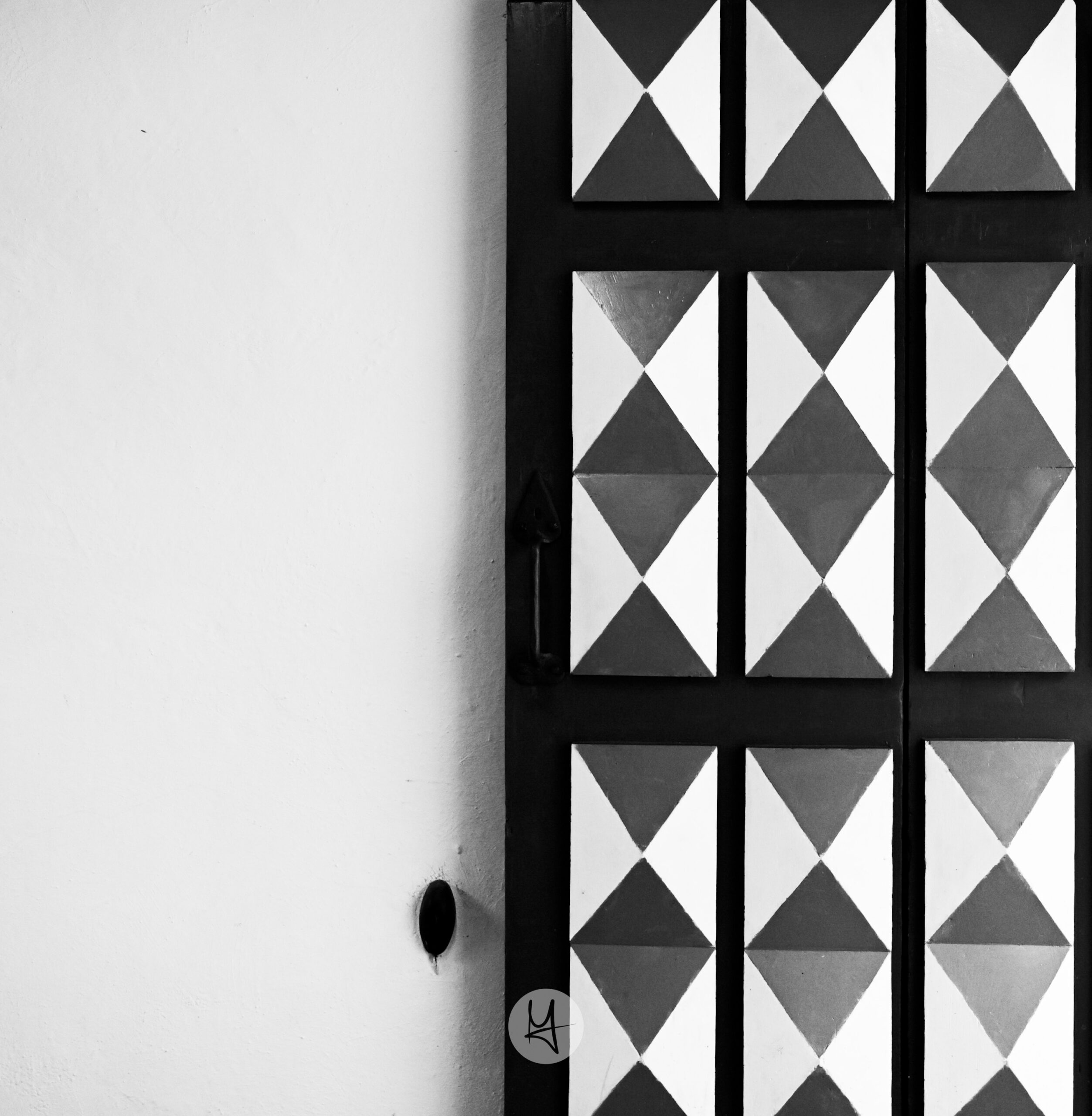 Black and white photograph of a detailed door in Tallinn, Estonia, which takes half of the image. The door is constructed of dark wood panels, each divided into a triangular pattern of black and white. The pattern repeats across the door. The background is a smooth, pale grey wall, with a strong side light creating a dramatic shadow.