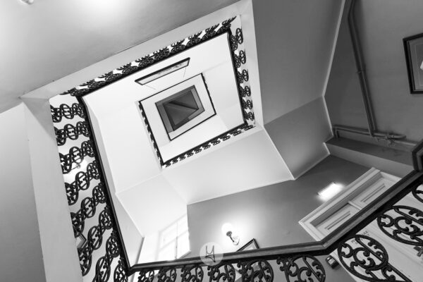 A black and white photo of a staircase in an old building in Tallinn. Looking from the bottom floor up to the top floor. The handrail spirals in a rectangular shape.