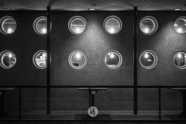 A black and white photo of a fassade of a restaurant. 12 small and round windows let you peek inside. In front of the fassade there is a empty small table.