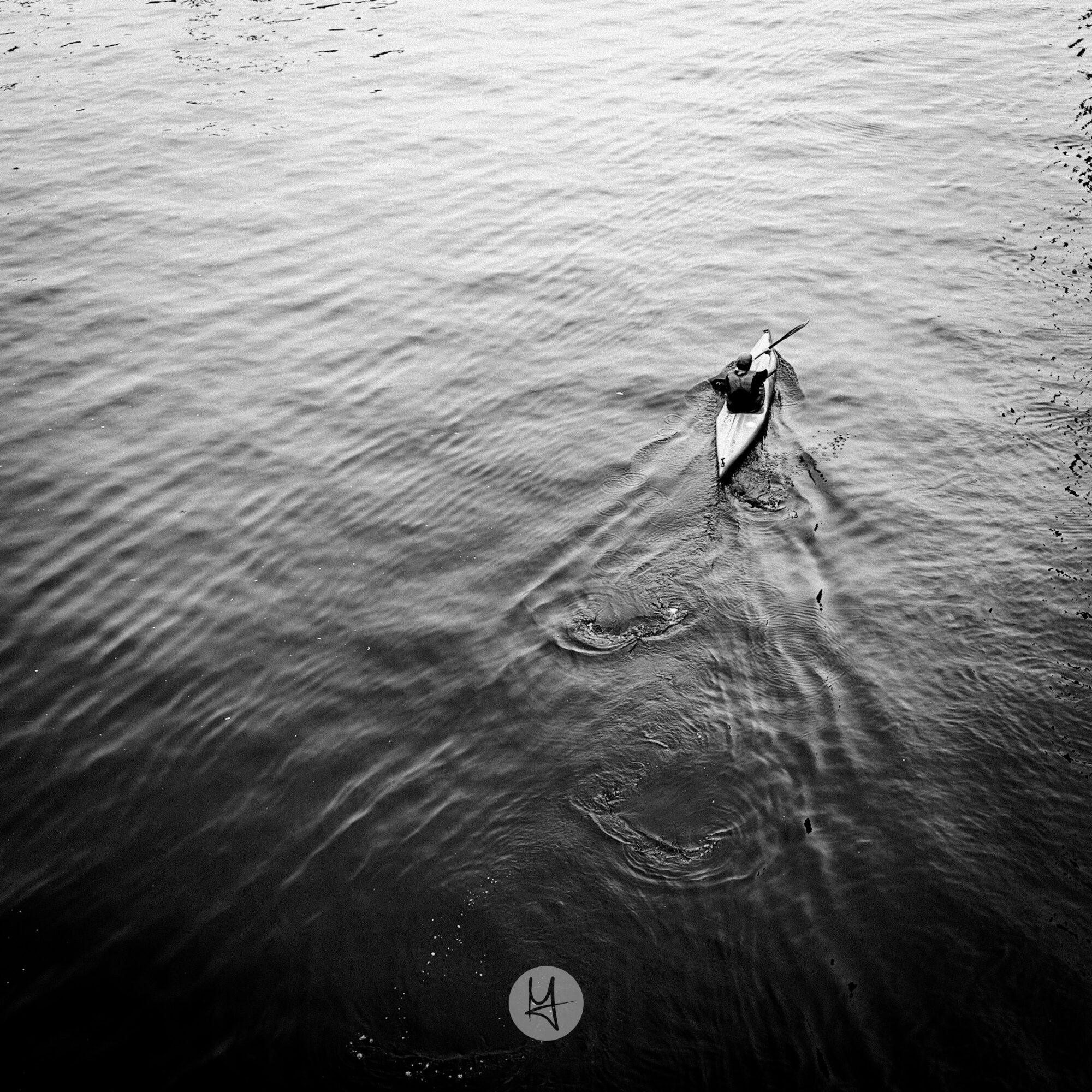 A high-angle, black and white shot of a person paddling a kayak on a river. The water is rippling, and the kayak is a narrow, streamlined shape.