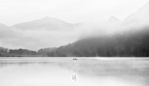 A view on the Haldensee lake in the Tannheimer Tal in Austria. The fog is covering most of the lake and the mountains. The sun is breaking through the fog. There are two fishermen in a boat on the lake.