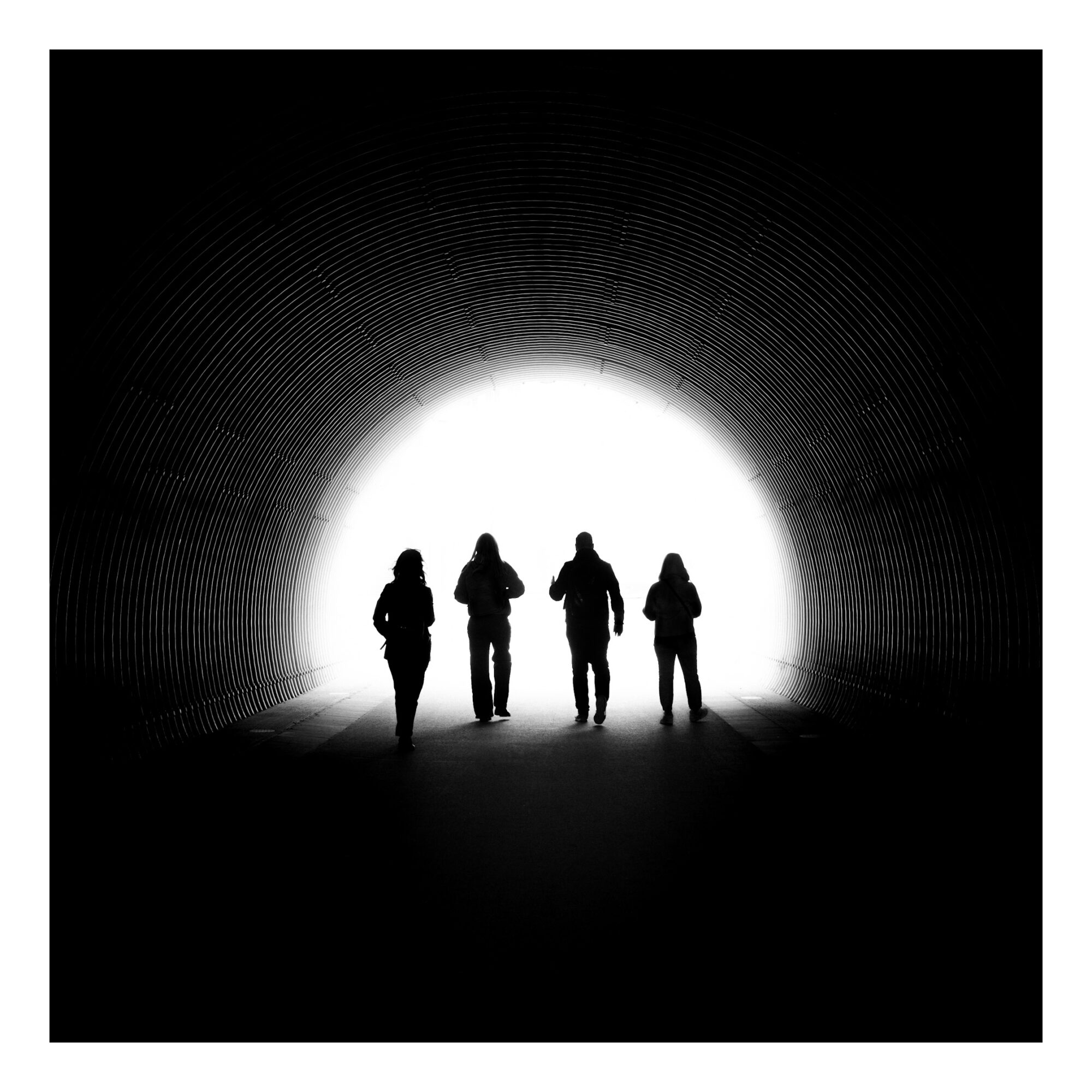 A high-contrast black and white photograph of four silhouetted figures walking through a cylindrical, corrugated metal tunnel. The concentric, rhythmic lines of the tunnel’s metal walls create a strong sense of perspective, drawing the eye toward the brilliant white light at the exit. The composition is symmetrical and dramatic, evoking a sense of journey, hope, and mystery.