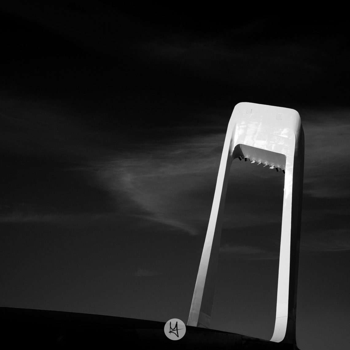 Black and white photo of a bridge in Tallinn. Only the middle column is visible and it’s shaped like a big upside down „u“. The background is dark with some feather clouds.