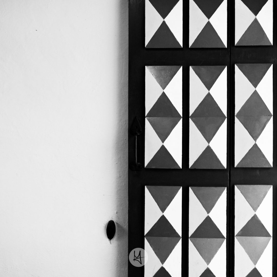 Black and white photograph of a detailed door in Tallinn, Estonia, which takes half of the image. The door is constructed of dark wood panels, each divided into a triangular pattern of black and white. The pattern repeats across the door. The background is a smooth, pale grey wall, with a strong side light creating a dramatic shadow.