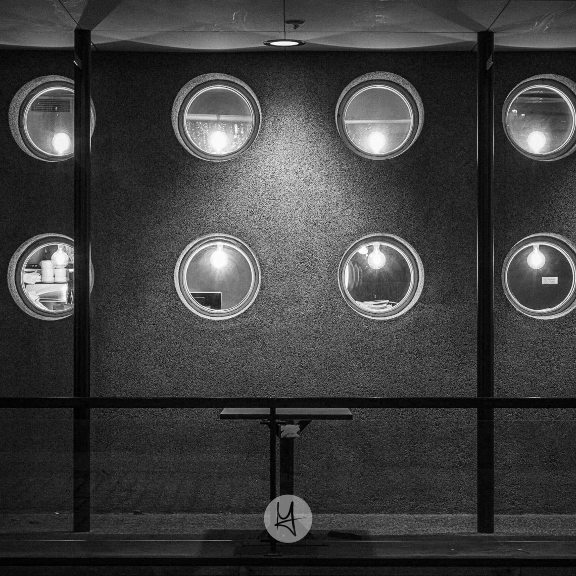 A black and white photo of a fassade of a restaurant. 12 small and round windows let you peek inside. In front of the fassade there is a empty small table.