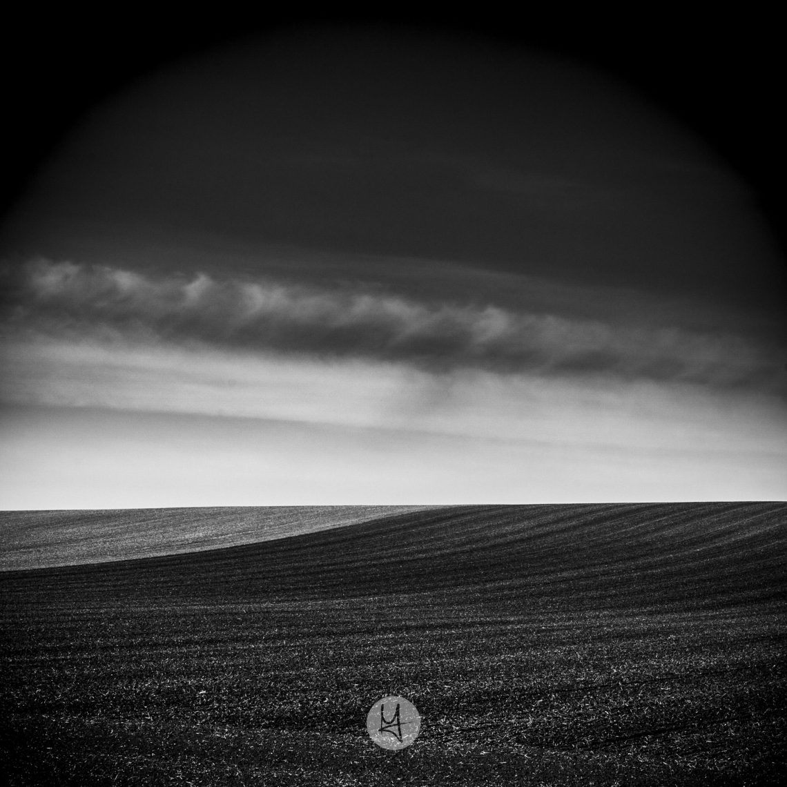 A black and white photo of a field. The foreground is a dark field with a faint pattern of furrows running from left to right, rising slightly to a curved horizon. A lighter band of sky sits above the horizon, with two distinct, dark cloud banks above it. The top of the frame is dark, with no detail visible.