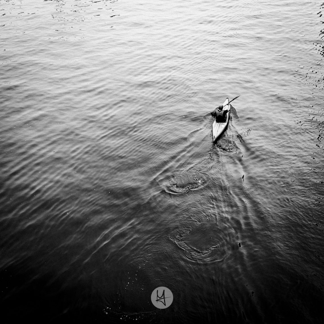 A high-angle, black and white shot of a person paddling a kayak on a river. The water is rippling, and the kayak is a narrow, streamlined shape.