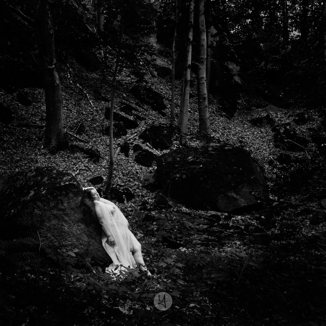 Ein monochromes, künstlerisches Foto, das eine nackte Frau in einem hellen, fließenden Schleier zeigt, die auf einem von Blättern bedeckten Felsen liegt. Die Person, halb im Profil, scheint in eine Pose der Ruhe oder des Schlafes versunken zu sein. Das Bild ist stark im Kontrast, mit dunklen Bäumen und Schatten im Hintergrund, die eine mysteriöse und etwas melancholische Atmosphäre schaffen.