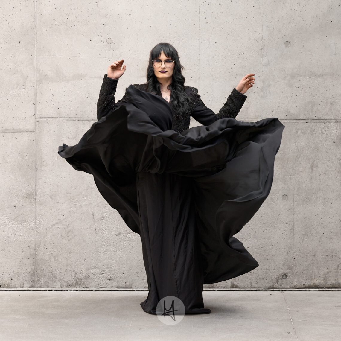 A portrait of a person with long dark hair and glasses, wearing a black top and a long black skirt. The skirt is caught in motion, billowing outwards. The person is standing in front of a concrete wall.